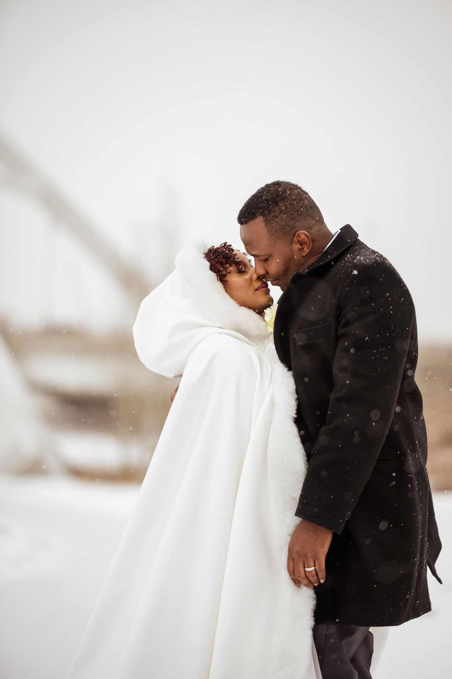 African American bride and groom share a romantic kiss in snowy winter wonderland, bride in white fur cape, groom in black coat.