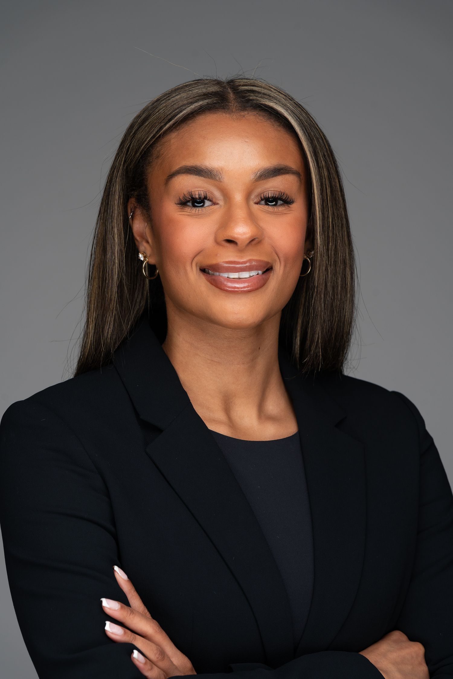 Professional headshot of someone in a black blazer and top smiling warmly with arms crossed against grey background.