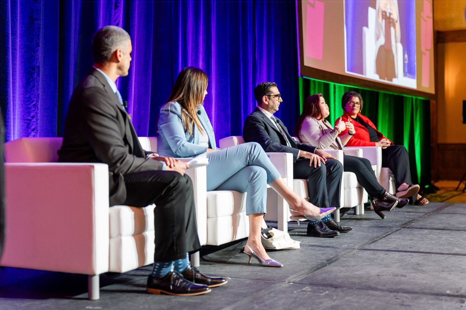 Panel discussion with speakers seated in white chairs on stage with blue curtain backdrop and presentation screen.