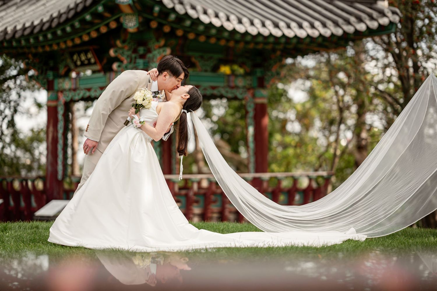 Couple sharing romantic kiss in white wedding dress and suit before traditional Asian pavilion outdoors.
