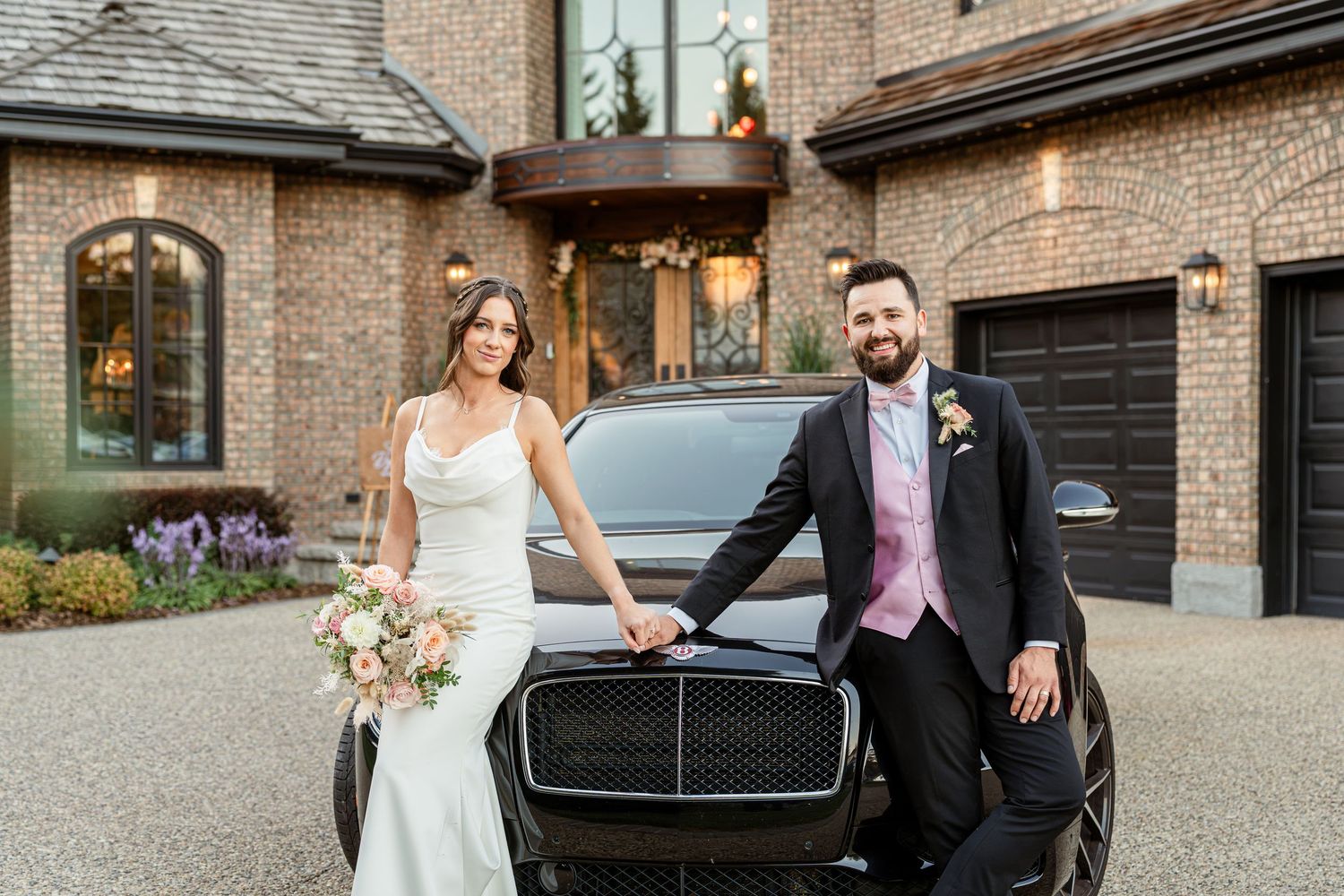 Bride and groom posing beside a luxury black SUV in front of an elegant brick mansion on their wedding day.