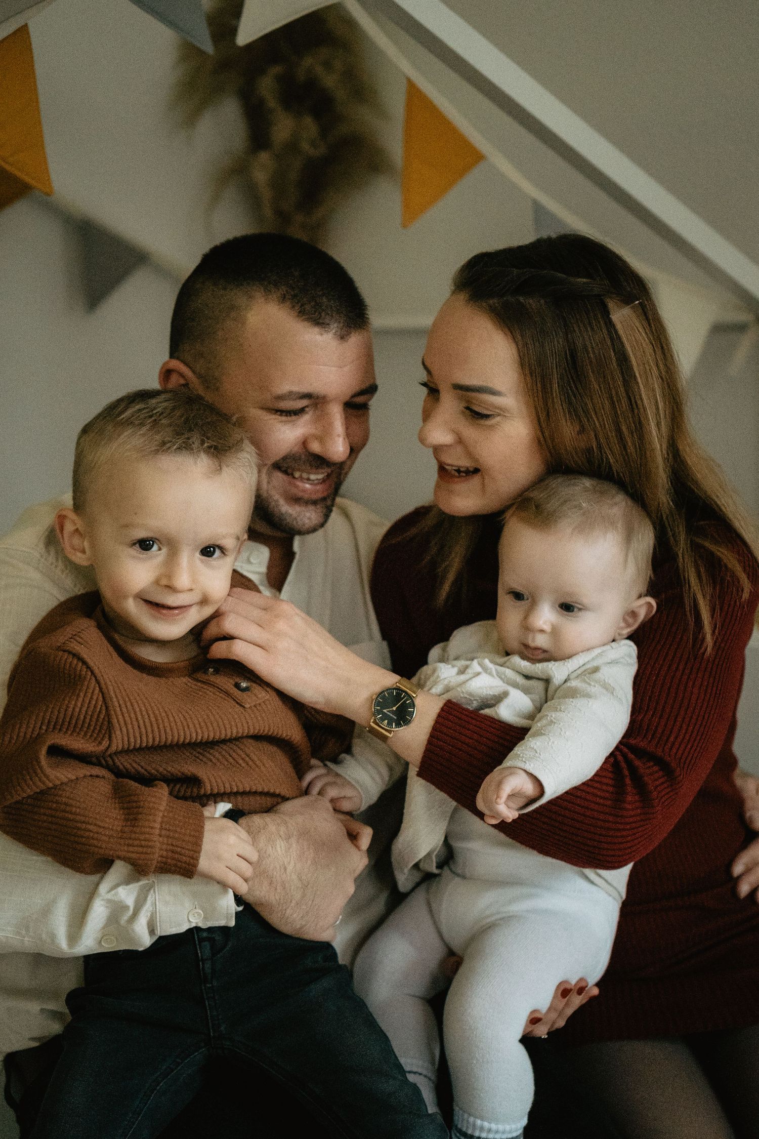 A warm family portrait indoors with triangle banners on the wall, showing parents and two young children in cozy sweaters.