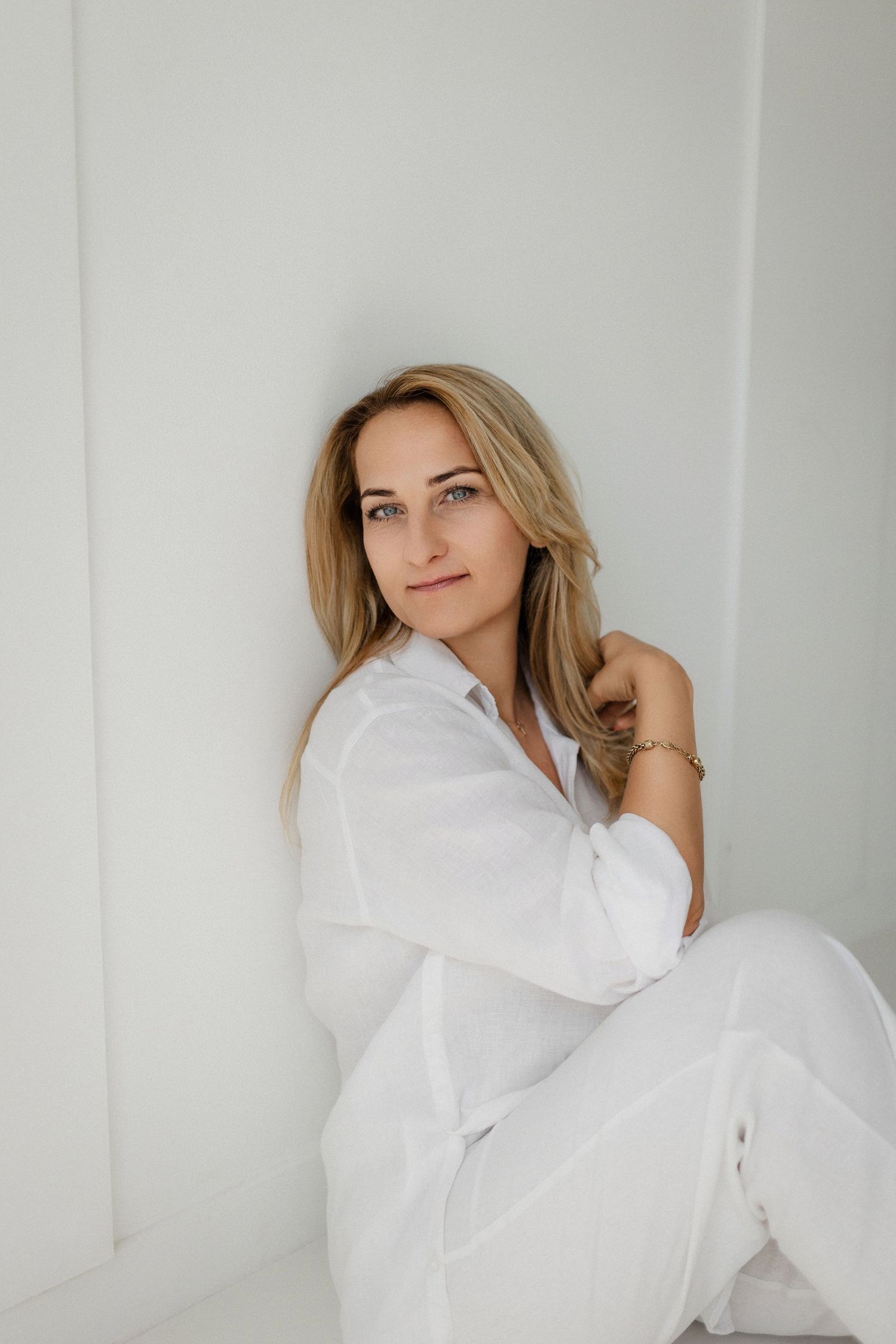 Woman in white clothing lays on light colored bedding while posing against white background for artistic portrait.