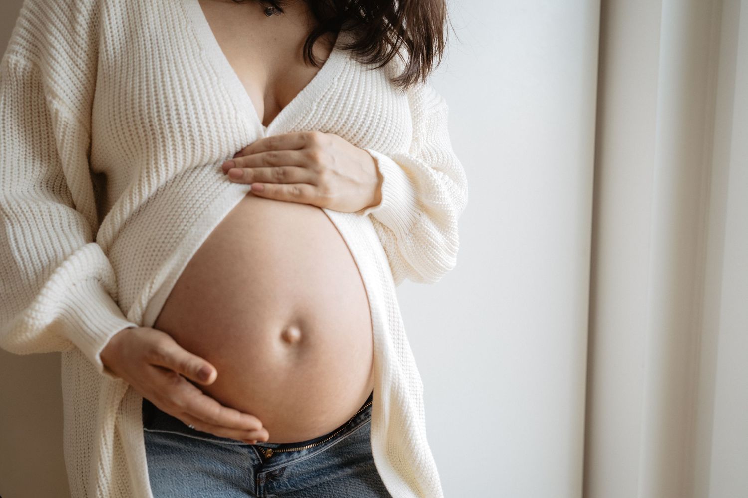 Pregnant woman in white cardigan holds her bare belly by window in soft natural light.