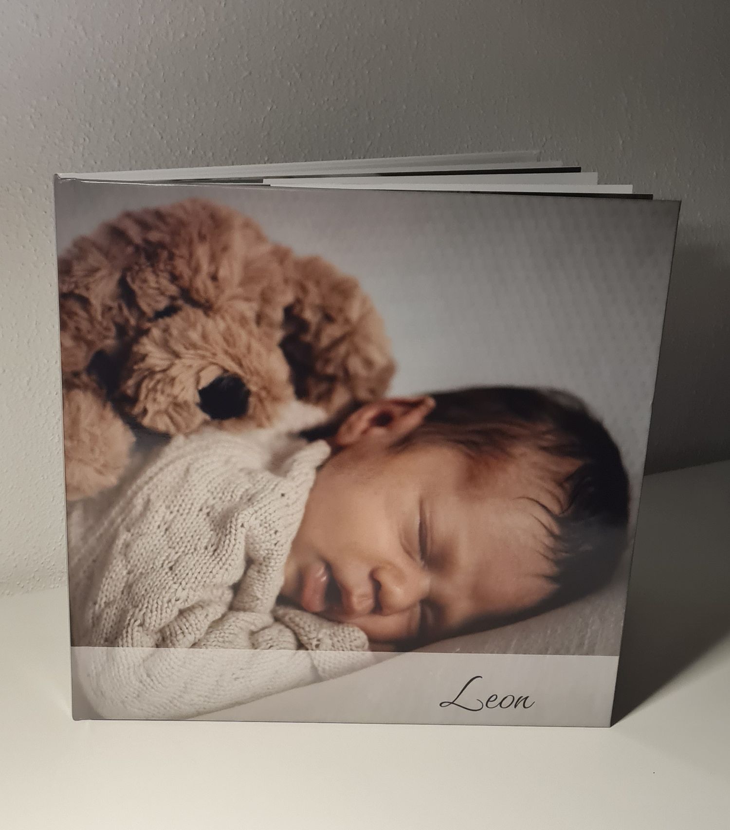 A photo album shows a sleeping baby in white knit clothing cuddled up with a soft brown teddy bear.