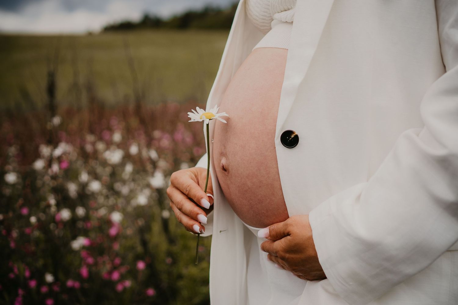 Person in a white dress holds their pregnant belly while standing in a field of pink wildflowers.