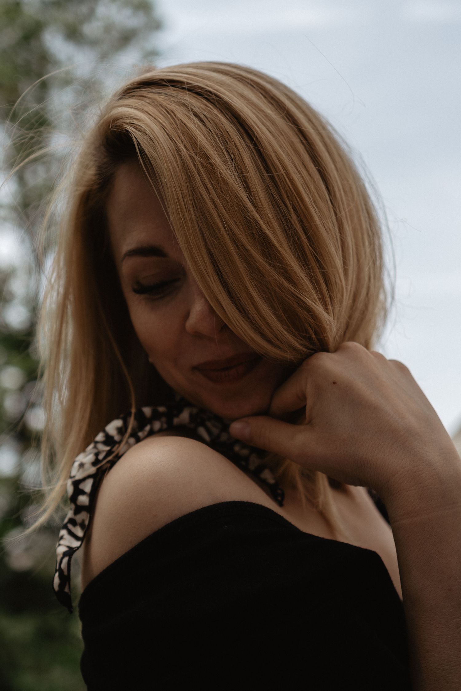 A person with honey blonde hair wearing a black sleeveless top stands in a thoughtful pose against a natural outdoor backdrop.