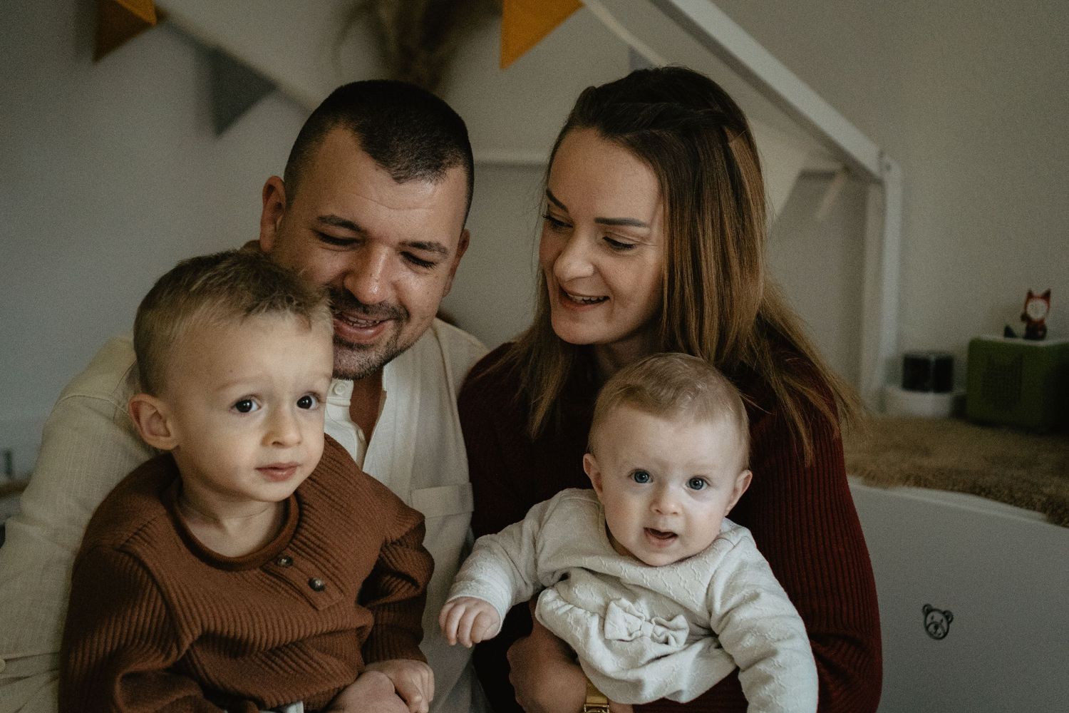 A family sitting together at home, with parents and two young children wearing cozy sweaters in warm lighting.