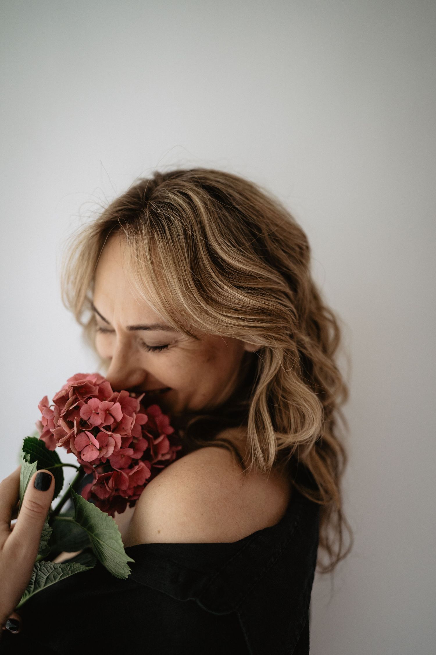 Woman in black top holding pink flowers against white wall while smiling with eyes closed.