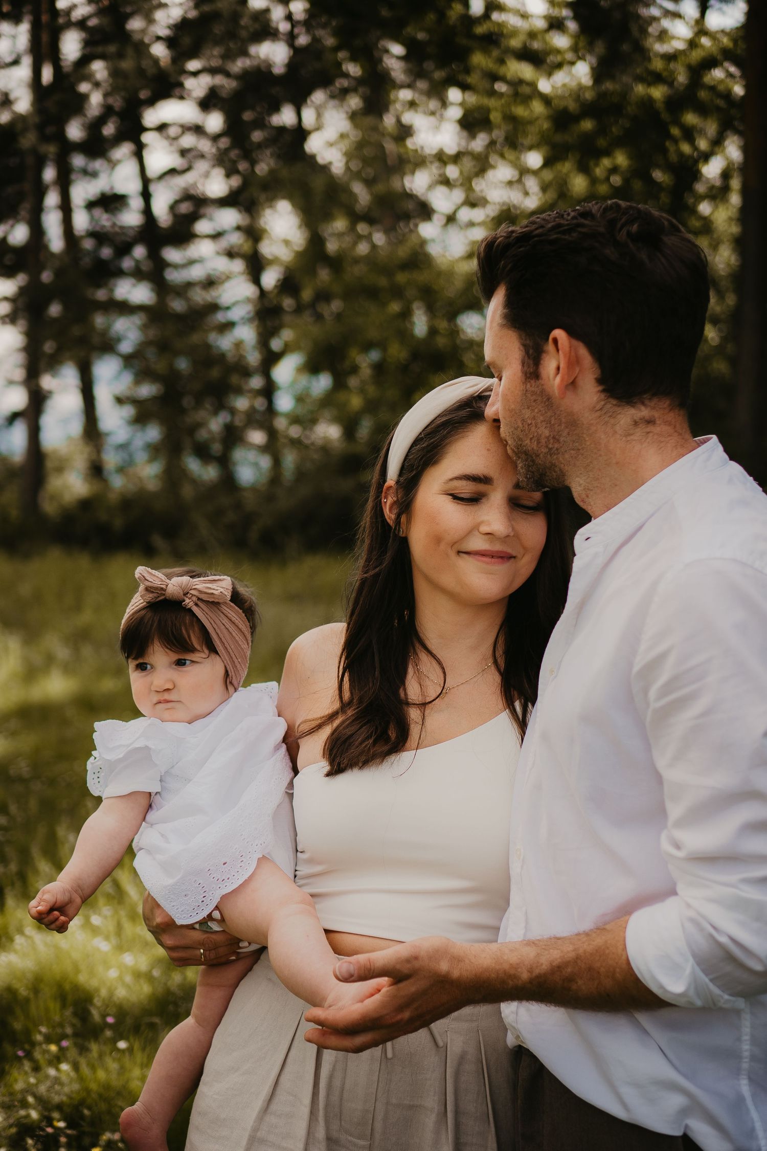 A tender family moment outdoors in warm sunlight, with a baby wearing a headband and white dress.