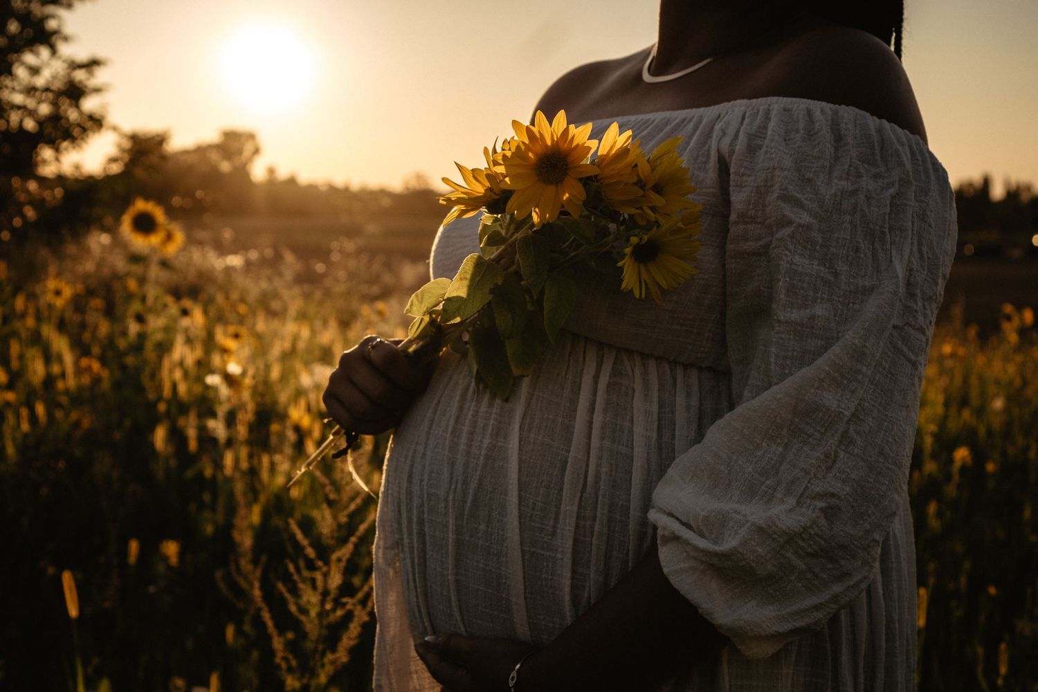 Silhouetted pregnant figure in white dress holds sunflowers against golden sunset in summer field.