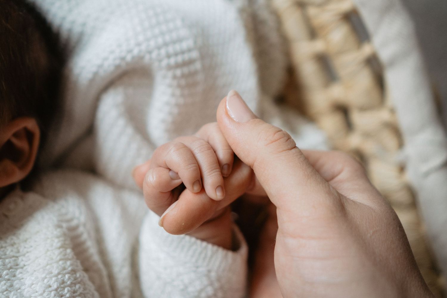 A tiny baby hand grasps an adult's finger while wrapped in a soft white knitted blanket.
