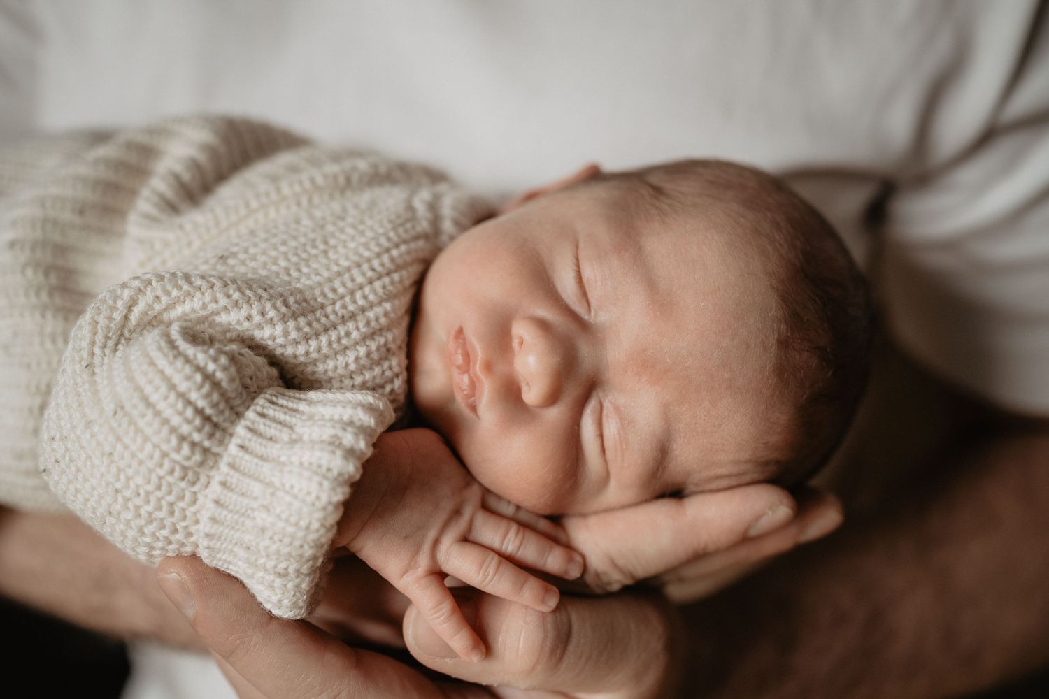 A newborn baby sleeps peacefully on white bedding while wearing a cozy knit sweater.
