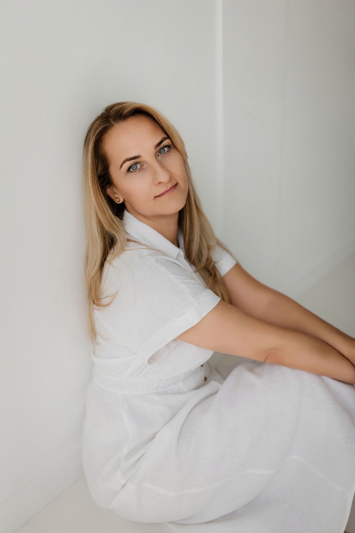 A person in a white dress lies gracefully against a white wall in an artistic pose.