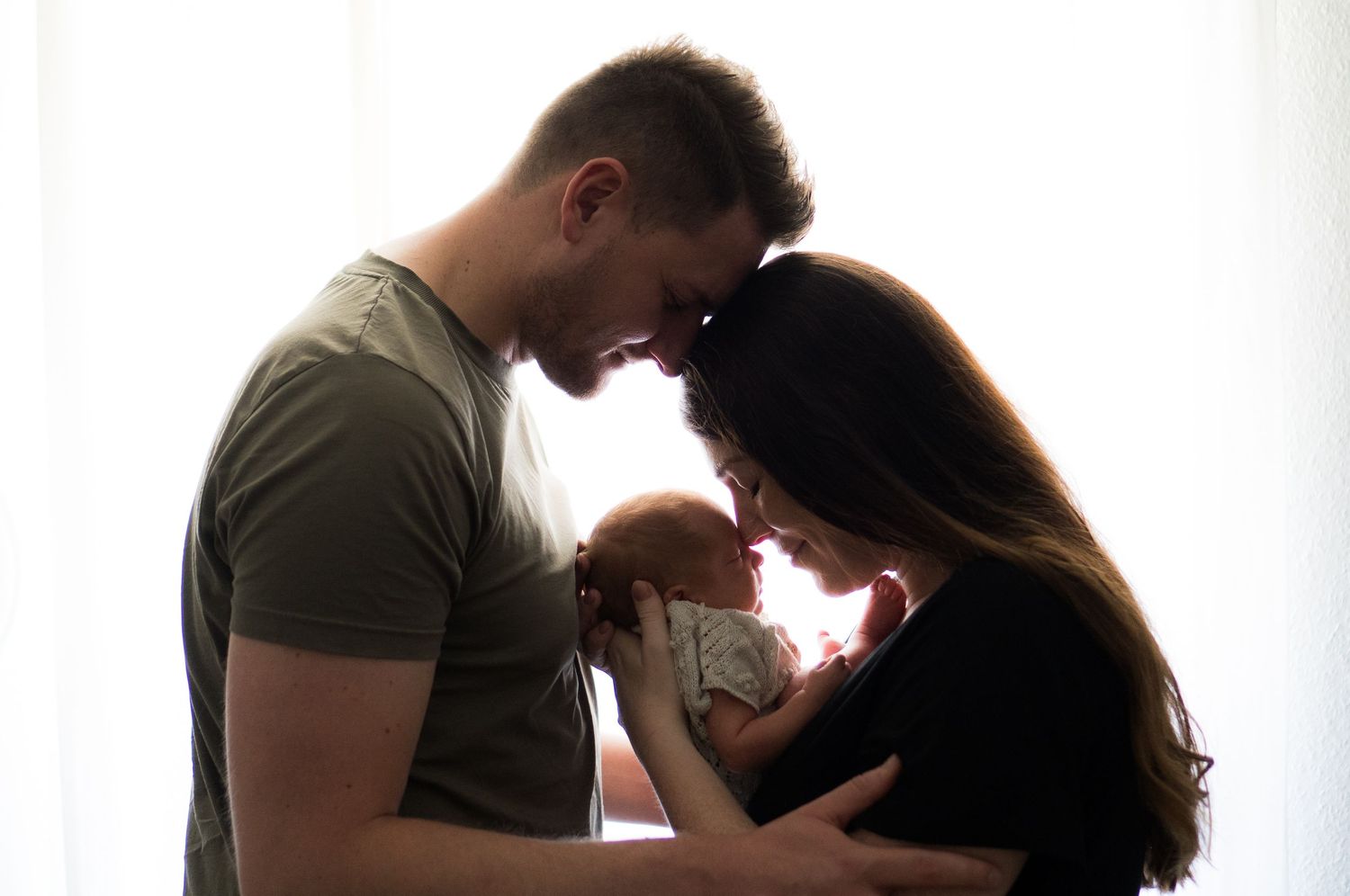 A silhouetted couple holds their newborn baby against a bright window, creating an intimate family portrait.