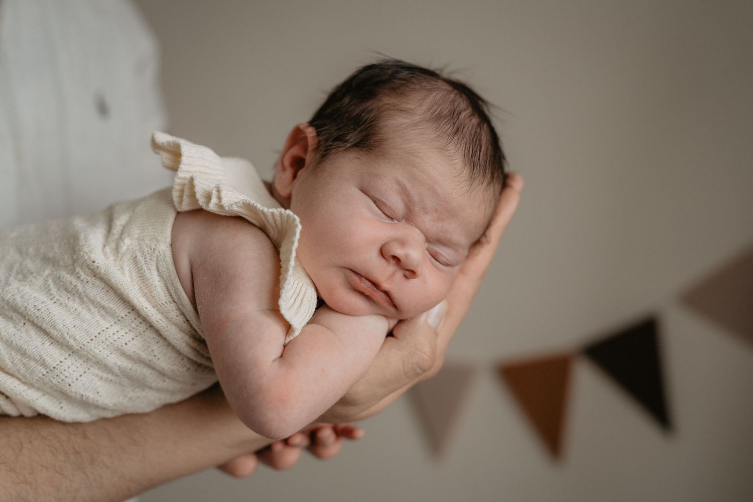 A sleeping newborn baby wrapped in white fabric is gently cradled in soft lighting against a neutral background.