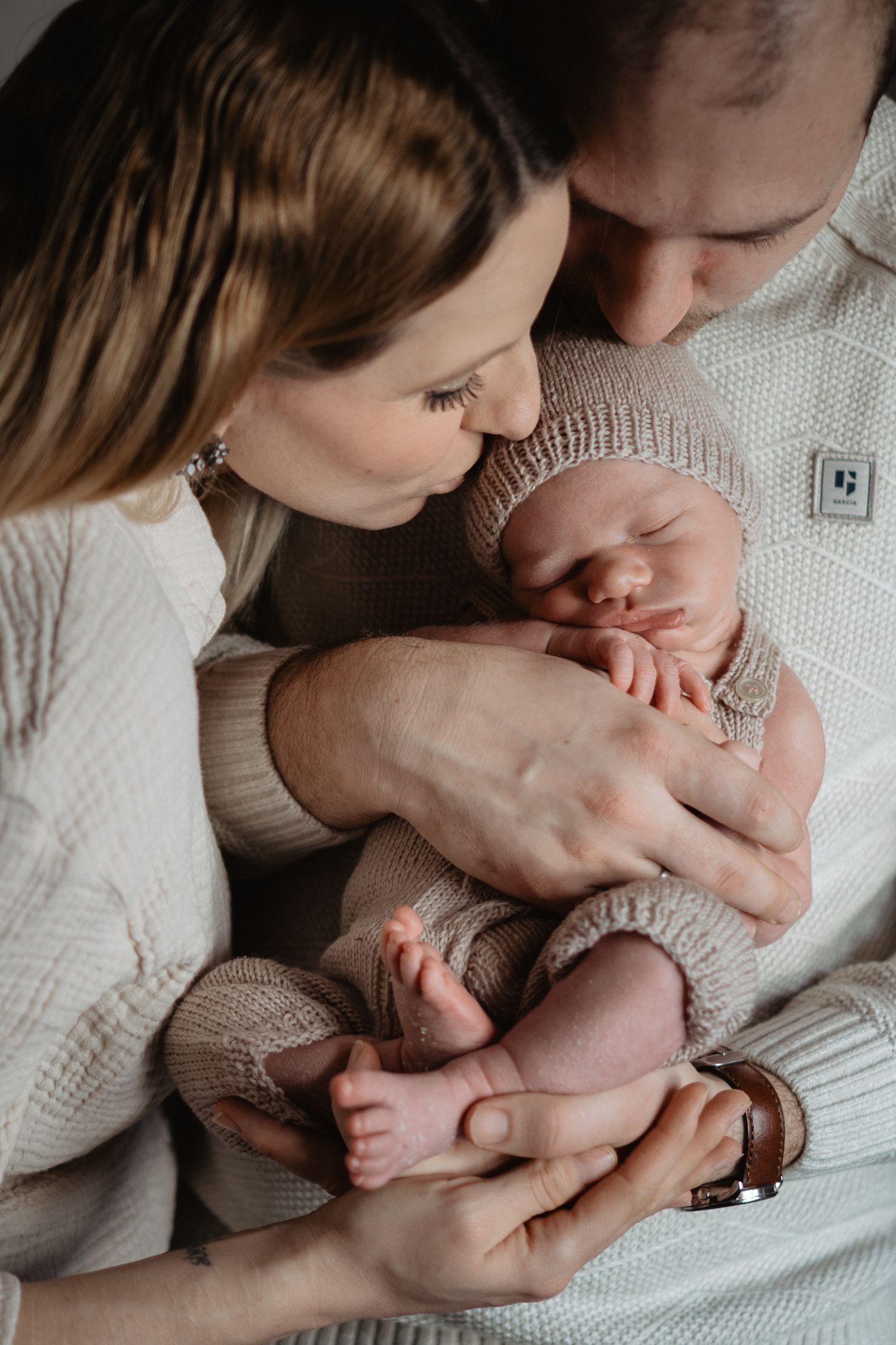 A sleeping newborn baby wearing a knitted beanie is tenderly held and gazed upon by parents in white sweaters.