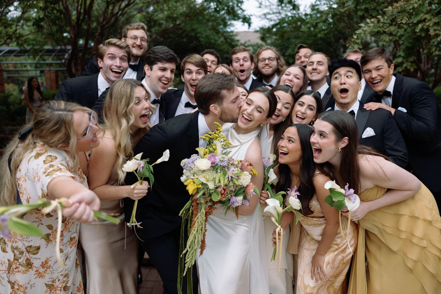 A joyous wedding party in formal attire laughs together outdoors while holding bouquets of yellow and white flowers.
