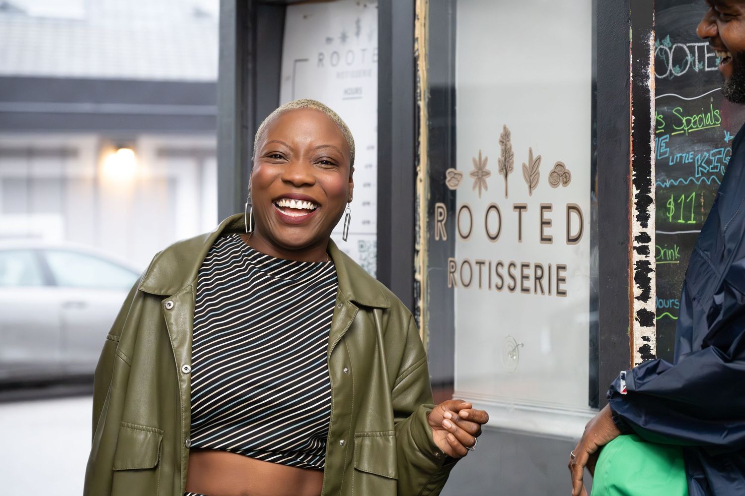 Woman smiling outside Rooted Rotisserie restaurant, wearing green leather jacket and striped crop top.
