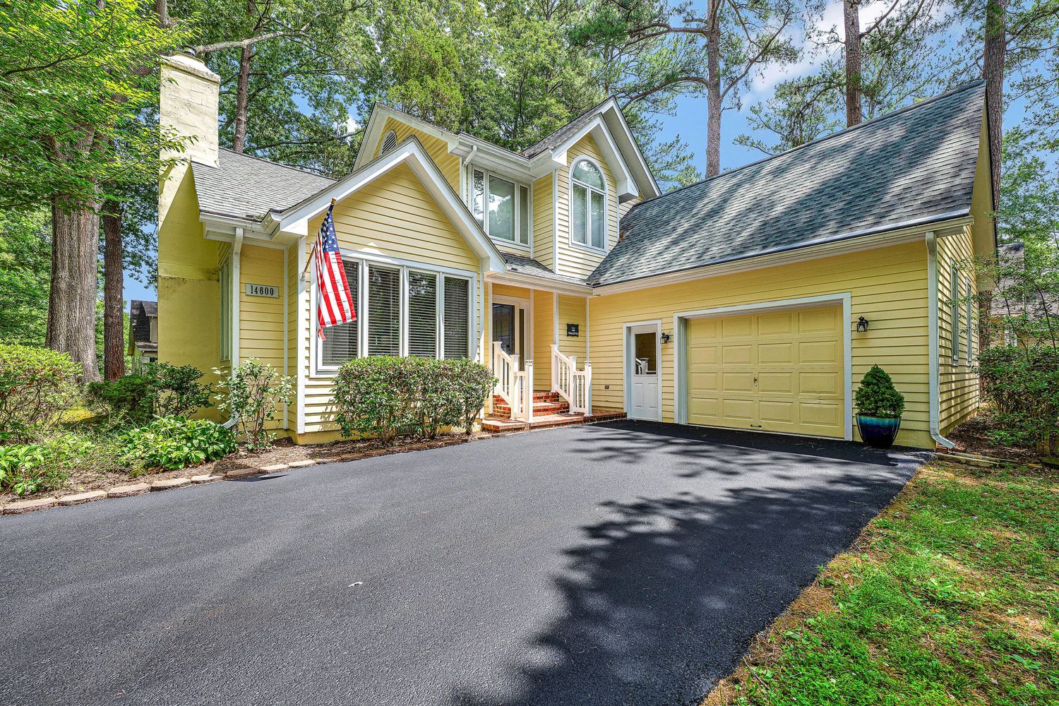 Yellow Cape Cod home with black driveway, attached garage, American flag, and lush green trees in background.