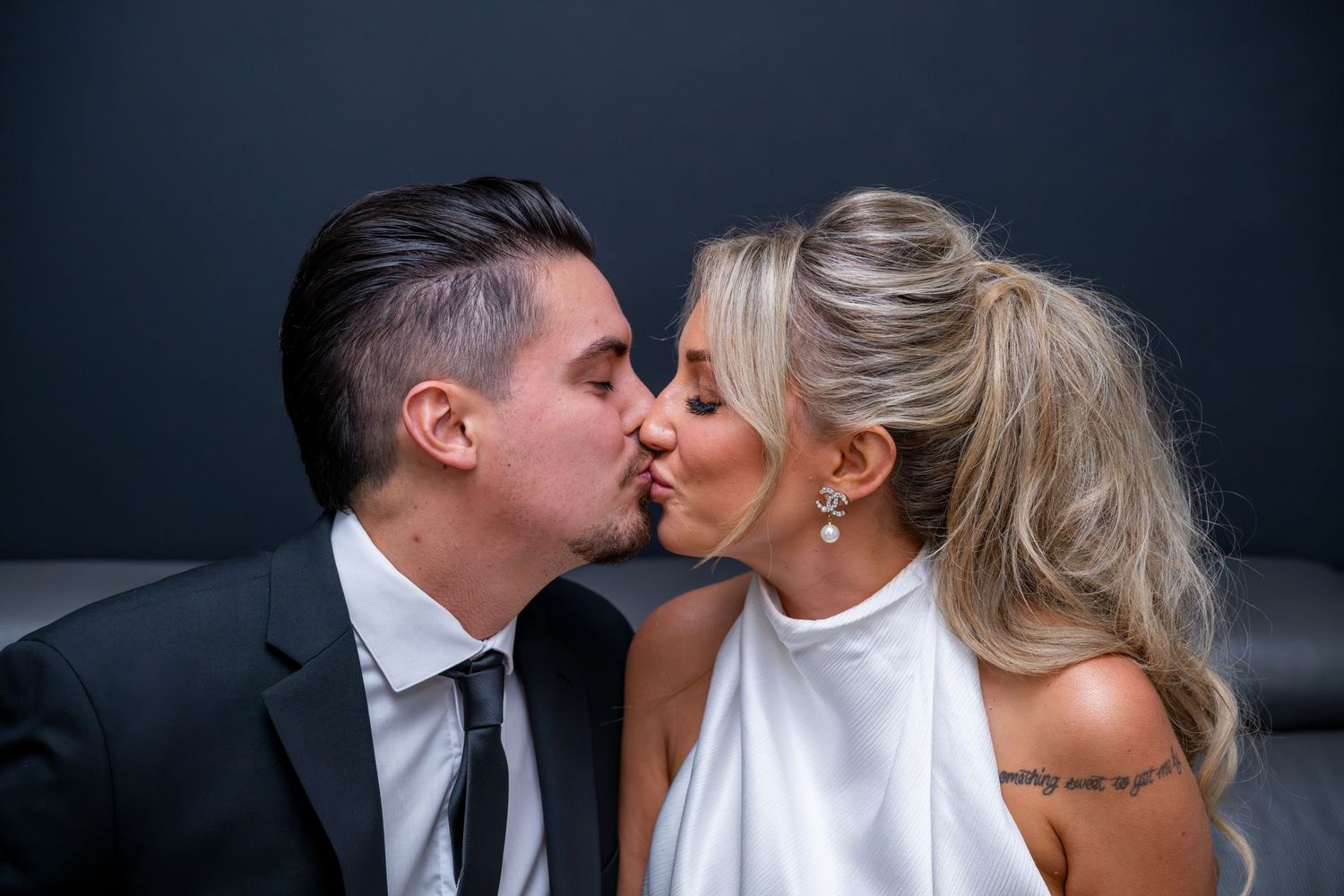 A couple shares a romantic kiss, the groom in a black suit and the bride in a white dress with blonde hair.