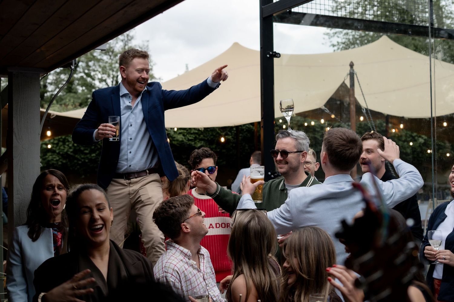 Wedding guest lifted up and pointing above a cheering crowd under a marquee