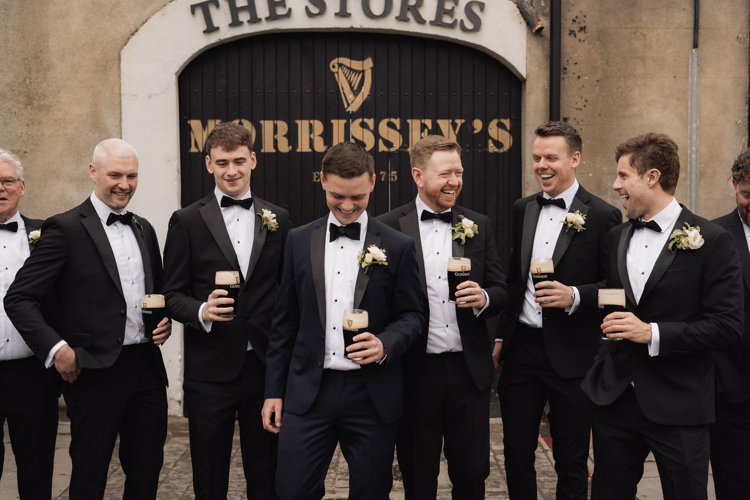 Groom and groomsmen in black tie holding pints outside a traditional pub before the wedding