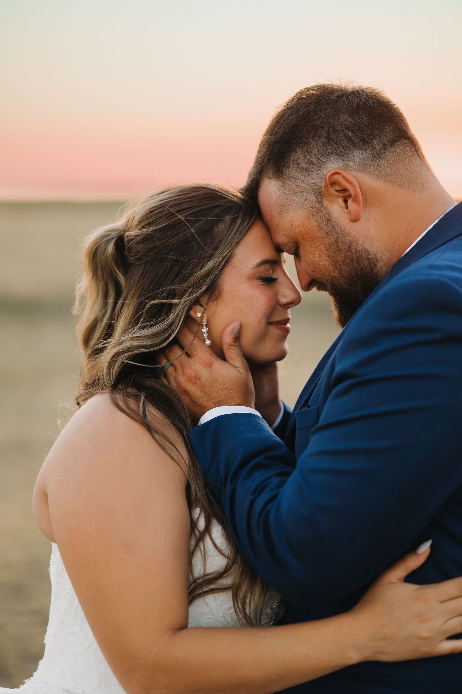 Bride and groom sharing an intimate forehead touch at sunset with a pink sky backdrop.