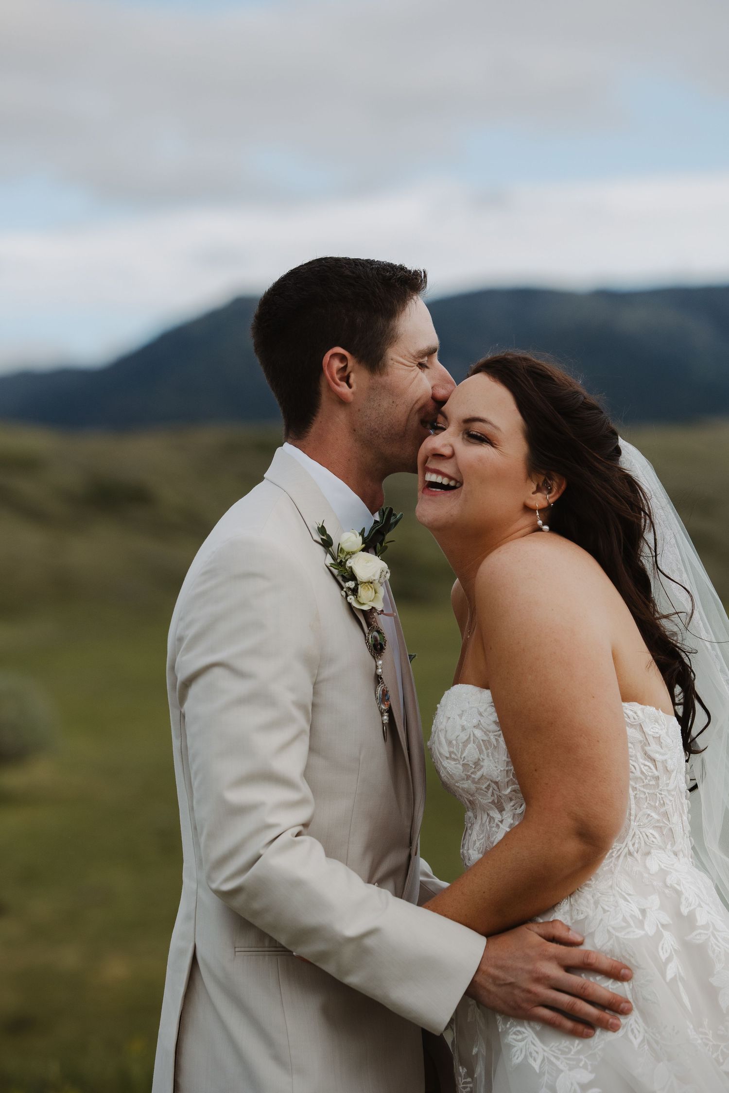 Laughing bride in strapless gown embraces groom in cream suit with mountain meadow backdrop.