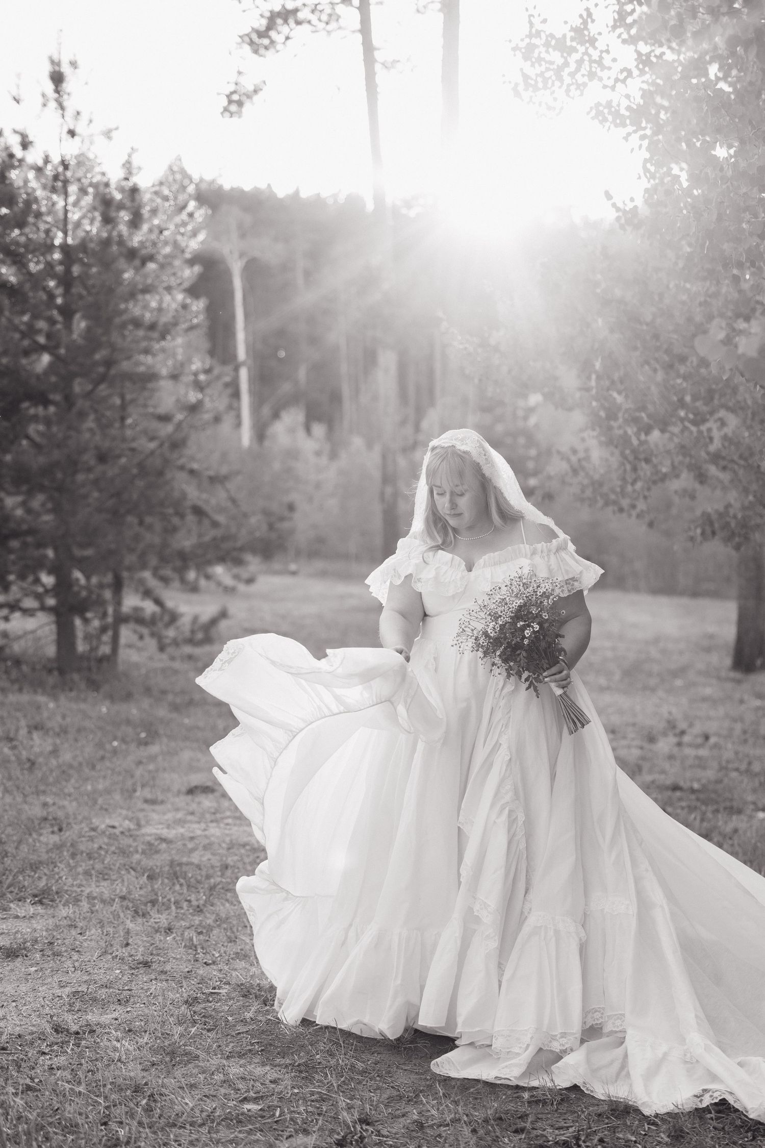 Black and white photo of bride in flowing off-shoulder gown with veil and bouquet in sunlit forest. Wyoming wedding.