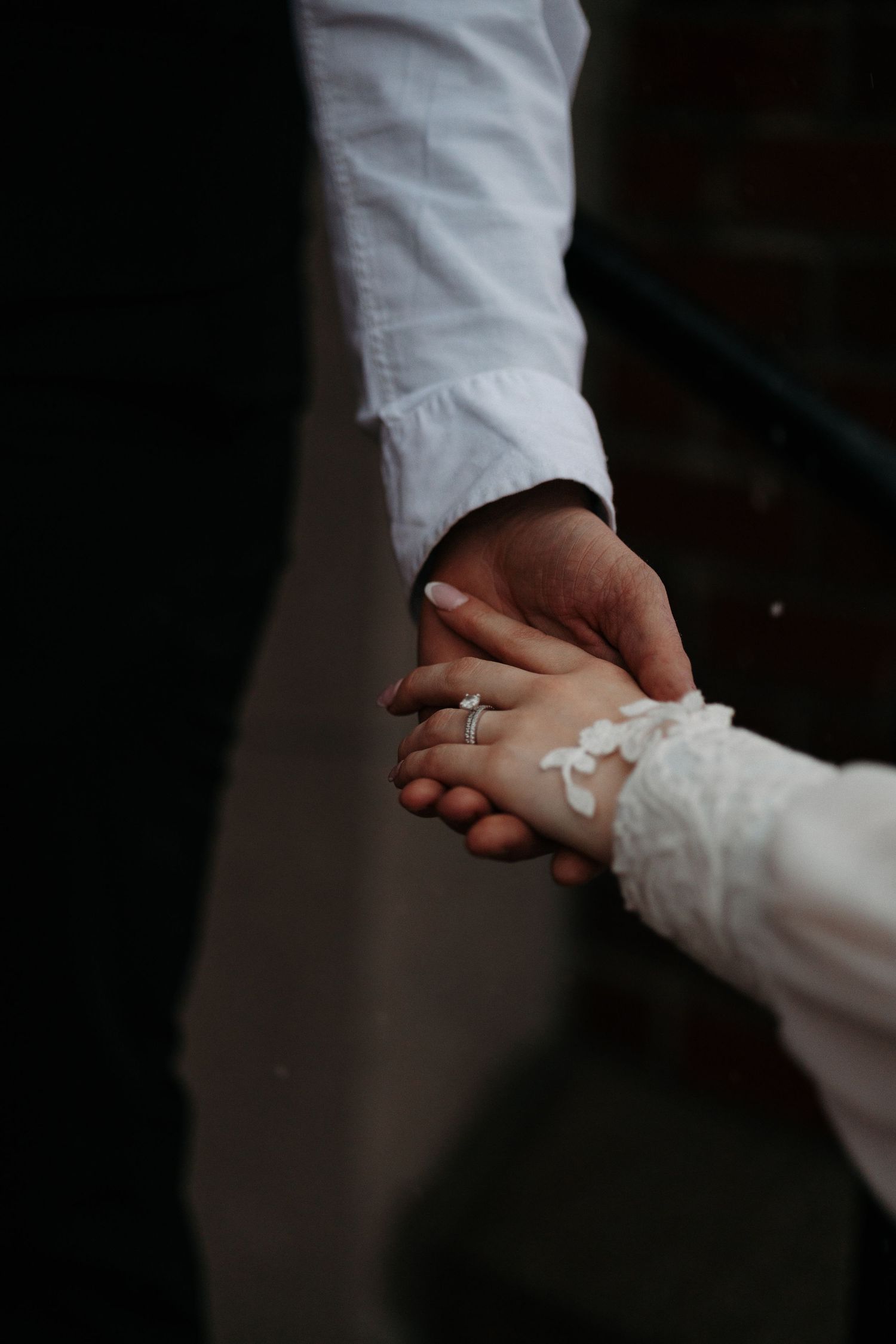 Close-up of bride and groom holding hands, showing engagement ring and lace wedding dress sleeve.