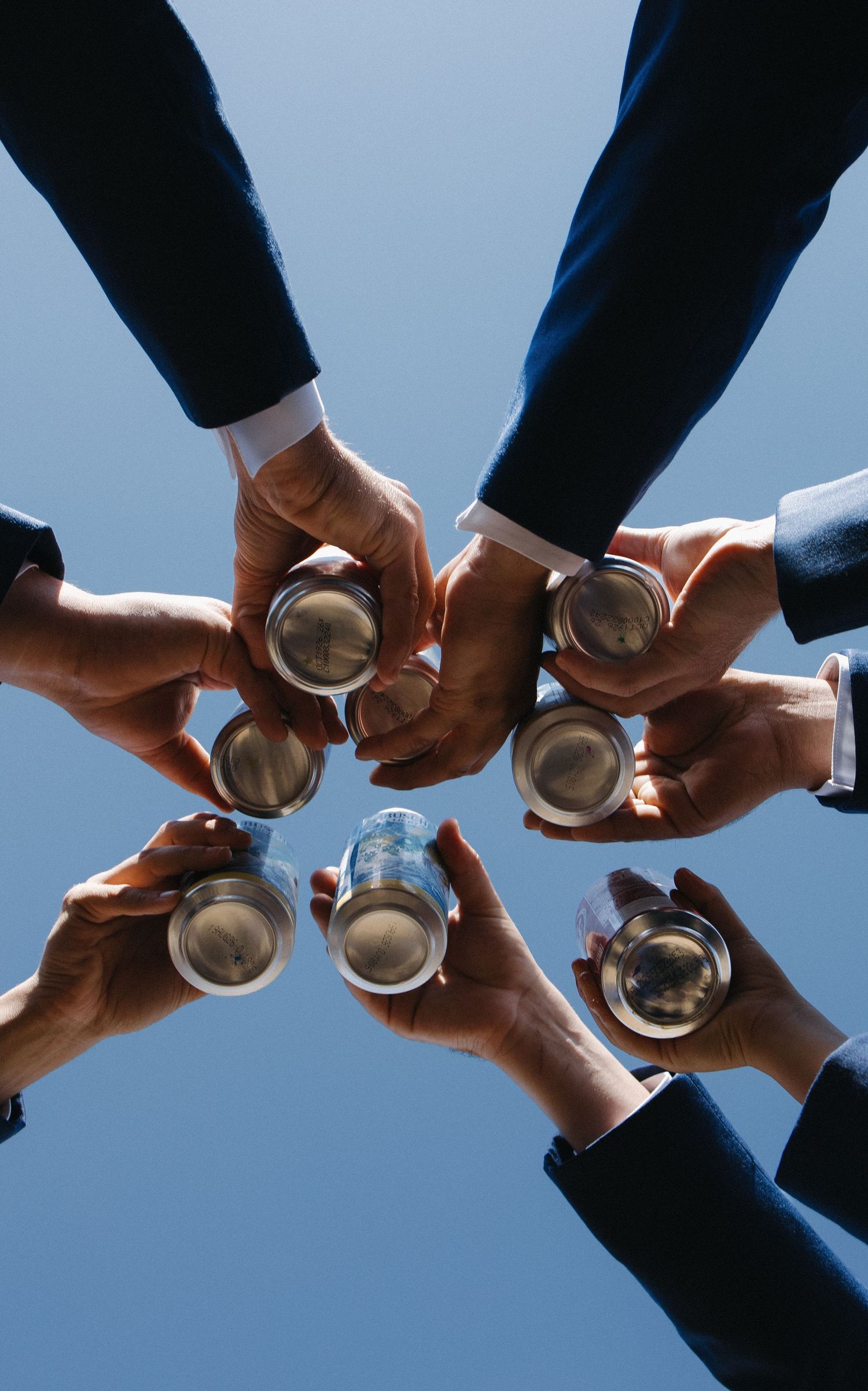 Low angle view of groomsmen in navy suits toasting with silver cups against a blue sky.