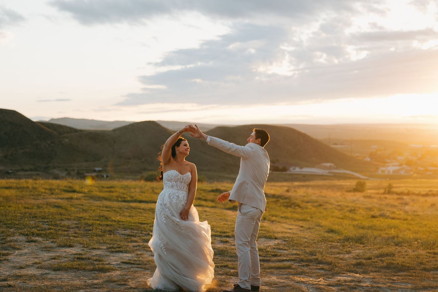 Groom twirls bride in layered white gown at golden sunset overlooking a scenic valley landscape.