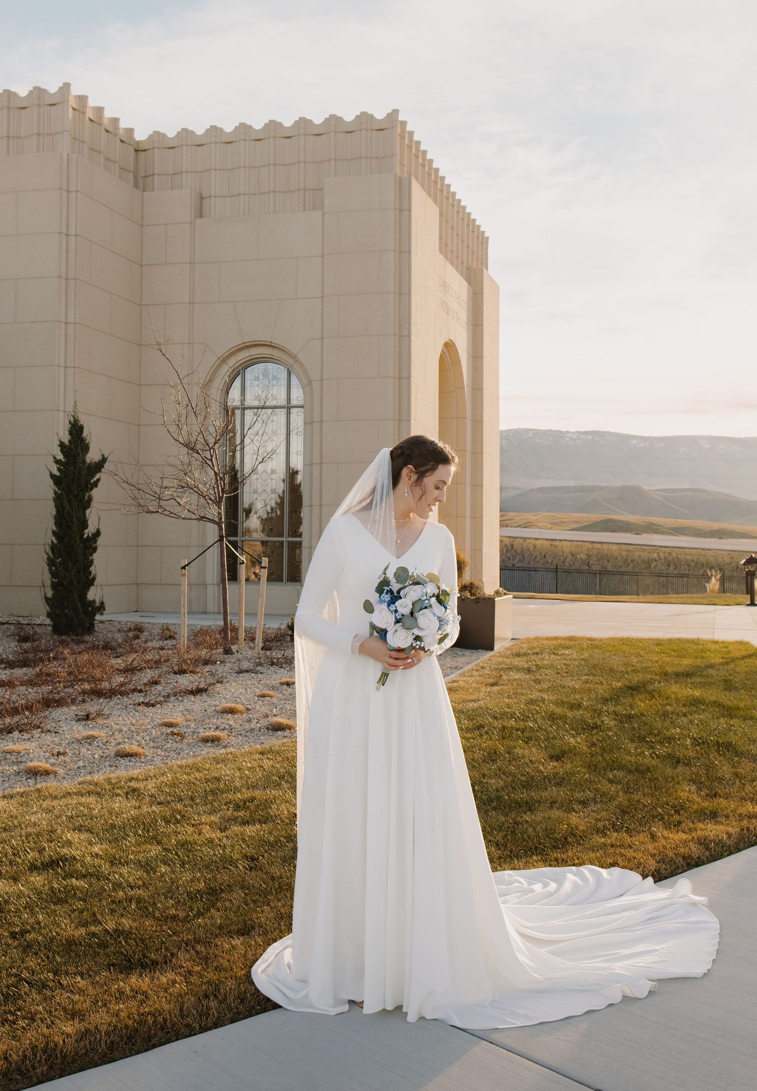 Bride in elegant long-sleeve white gown with veil holds bouquet outside a temple at golden hour.