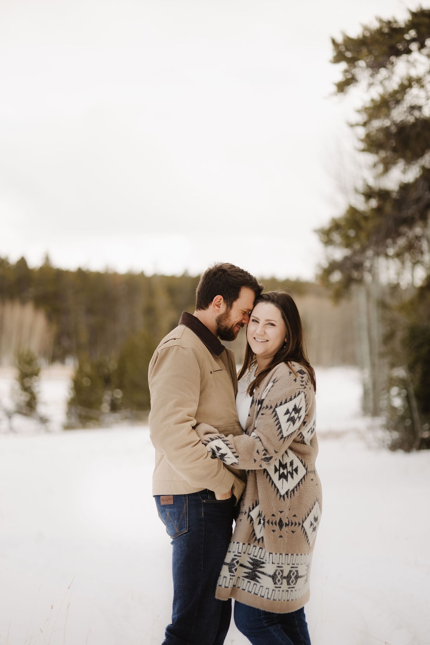 Couple embraces and smiles in a snowy winter forest wearing cozy neutral-toned outfits.
