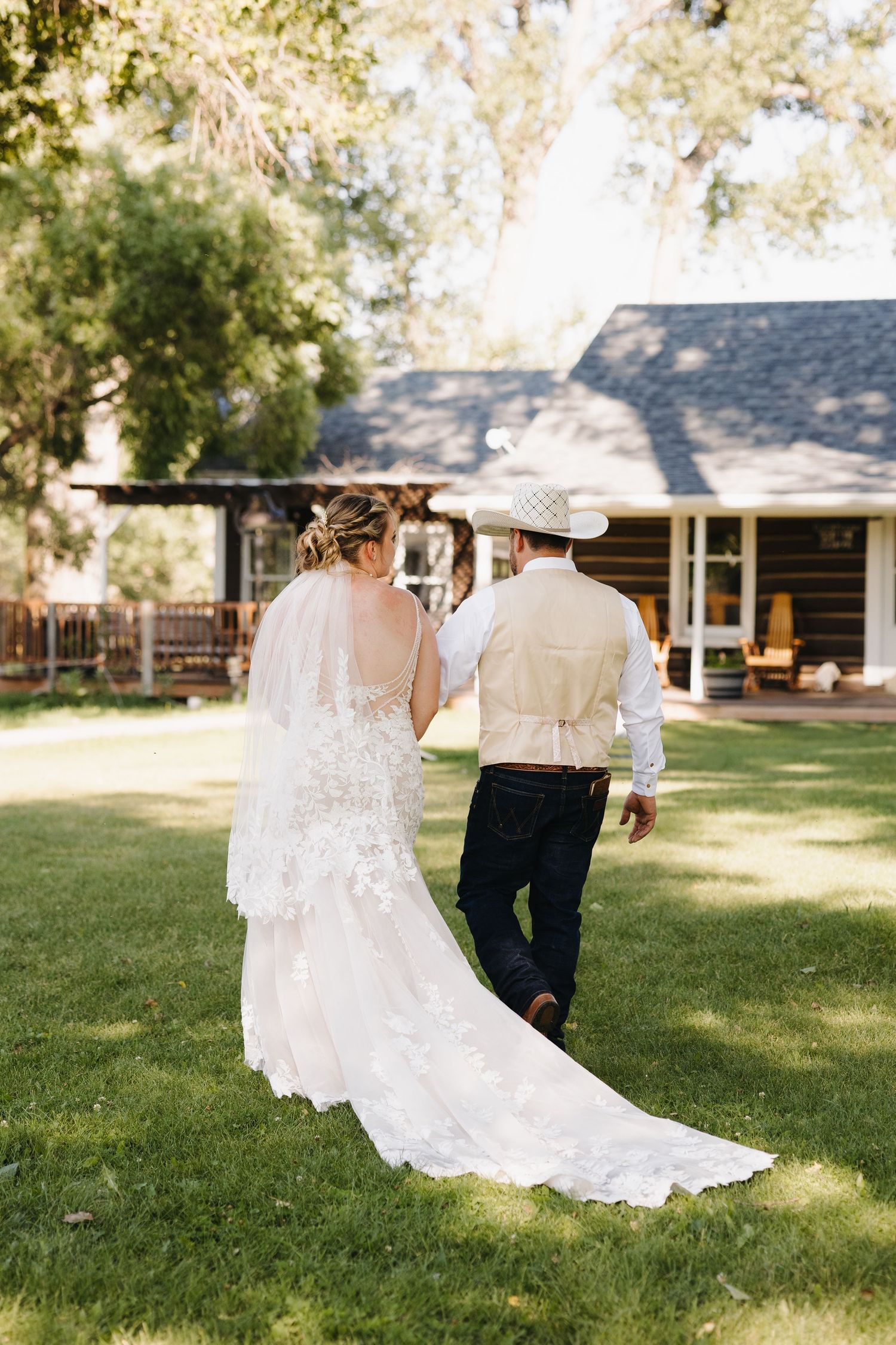 Bride in lace gown with veil walks arm-in-arm with groom in cowboy hat toward a rustic farmhouse.