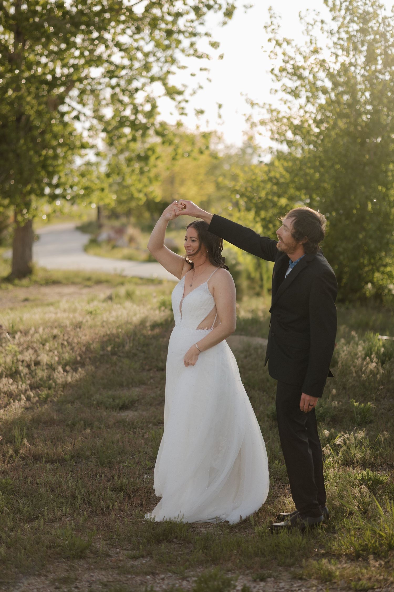 Groom twirling bride in white dress outdoors at golden hour among green trees.