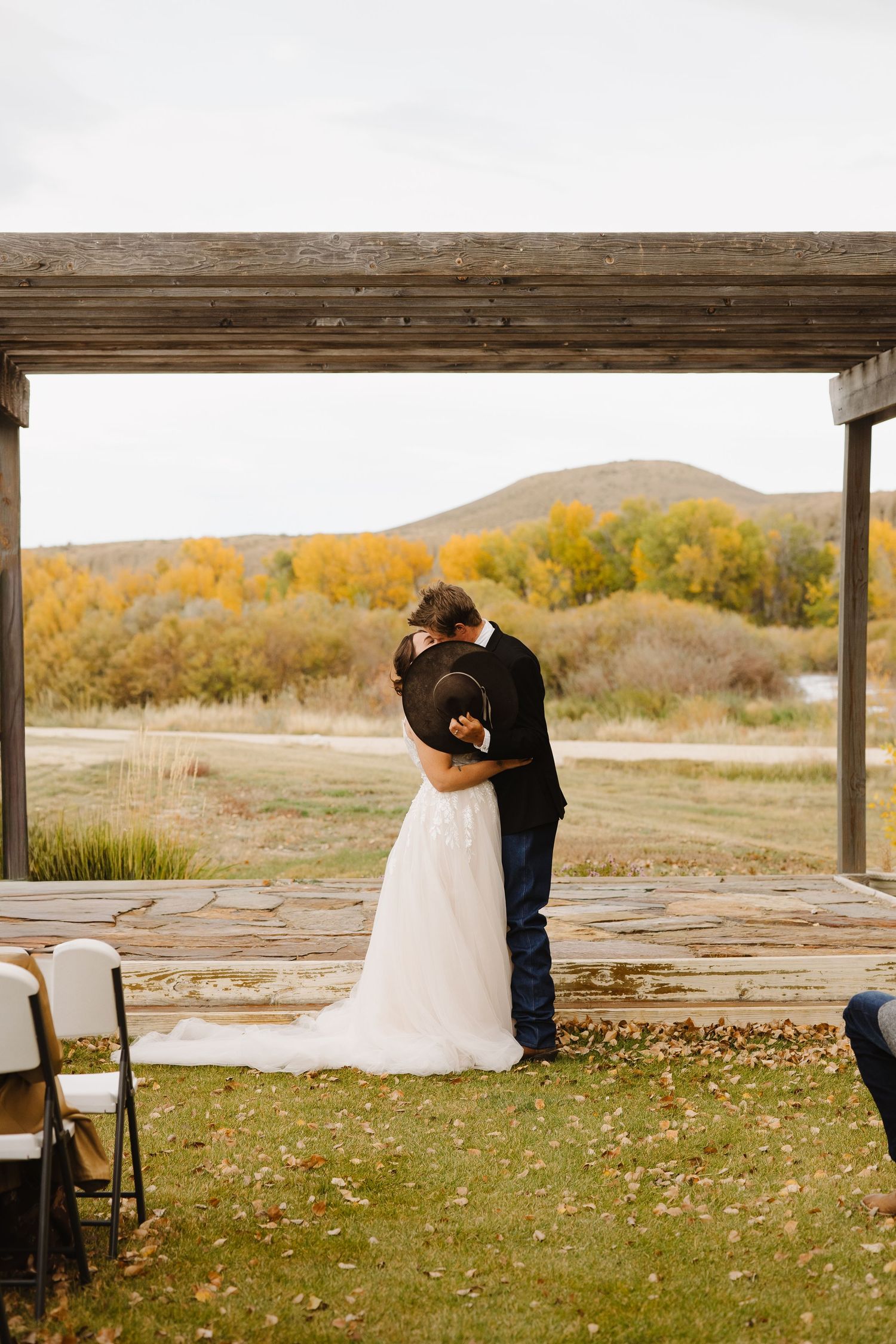 Bride and groom share first kiss under rustic wooden arch with autumn landscape backdrop.