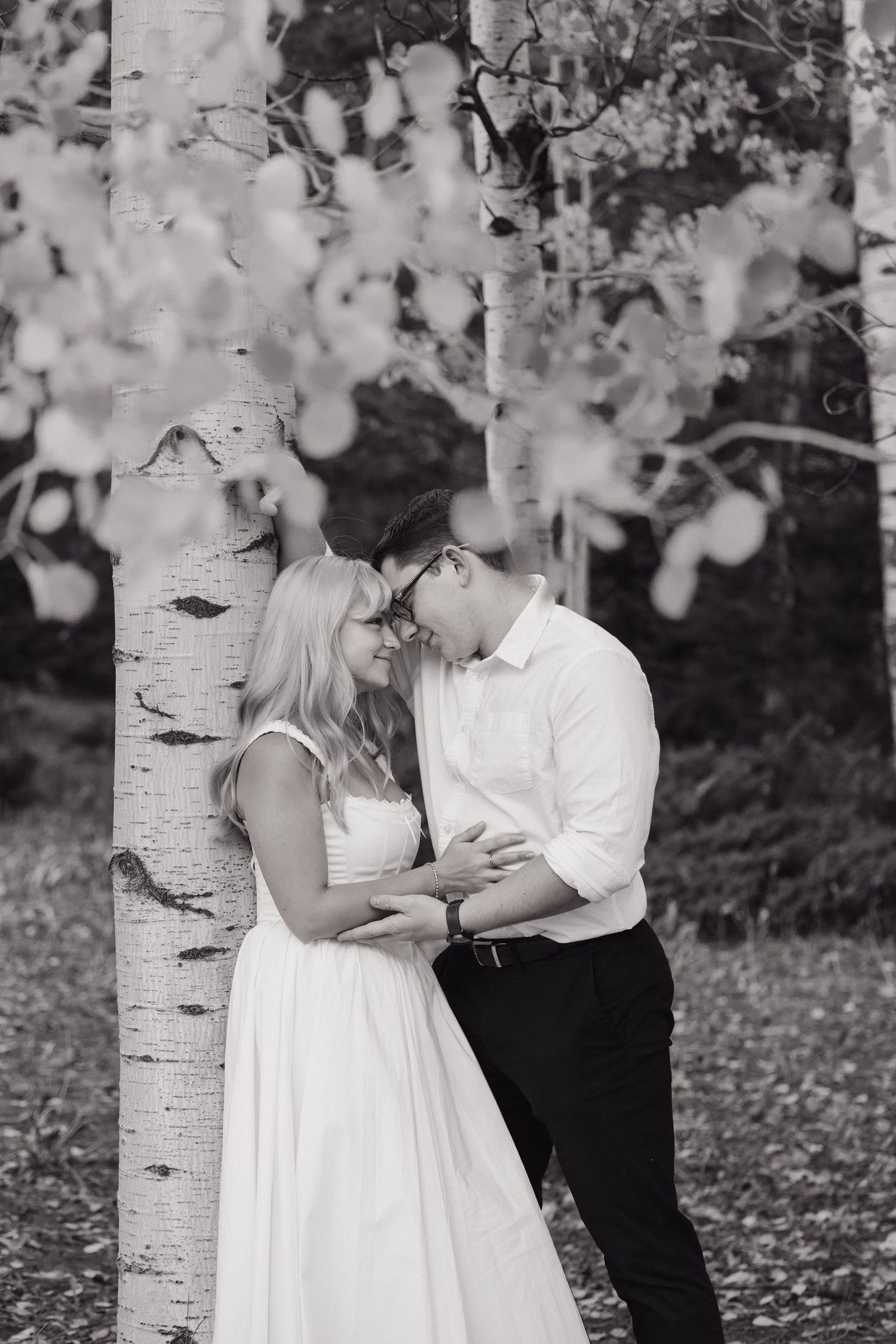 Black and white photo of couple sharing intimate moment leaning against white birch tree.
