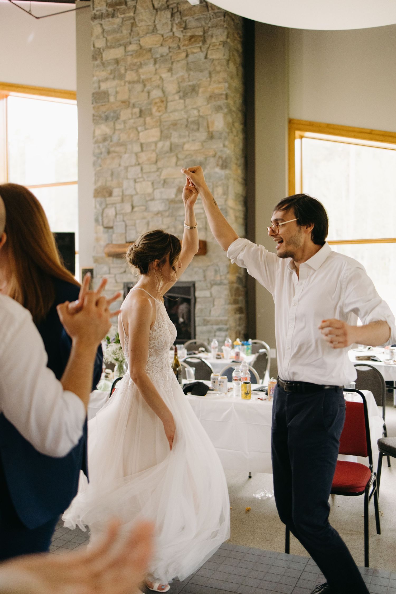 Bride in white tulle gown dances joyfully with guest near stone fireplace at wedding reception.