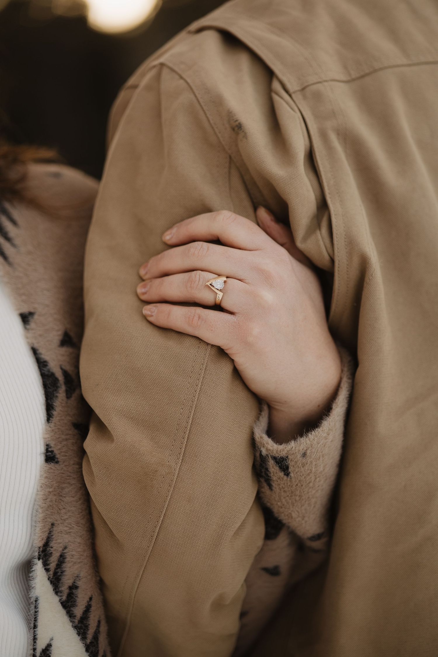 Close-up of a womans hand with a delicate engagement ring resting on a mans tan jacket. Wyoming engagement.