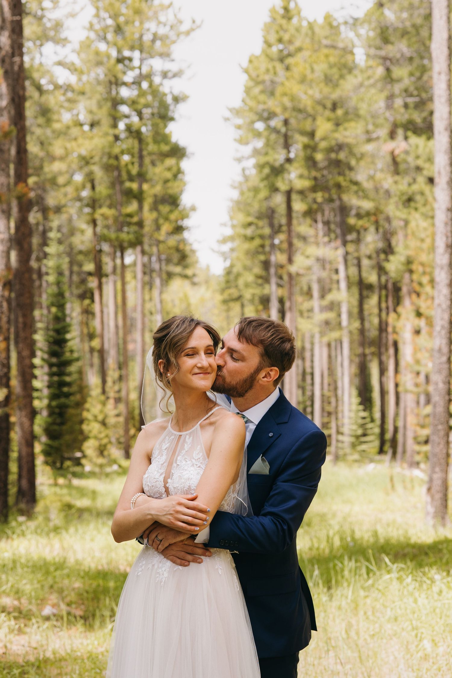 Groom in navy suit kissing bride on cheek while embracing her in a sunlit pine forest.