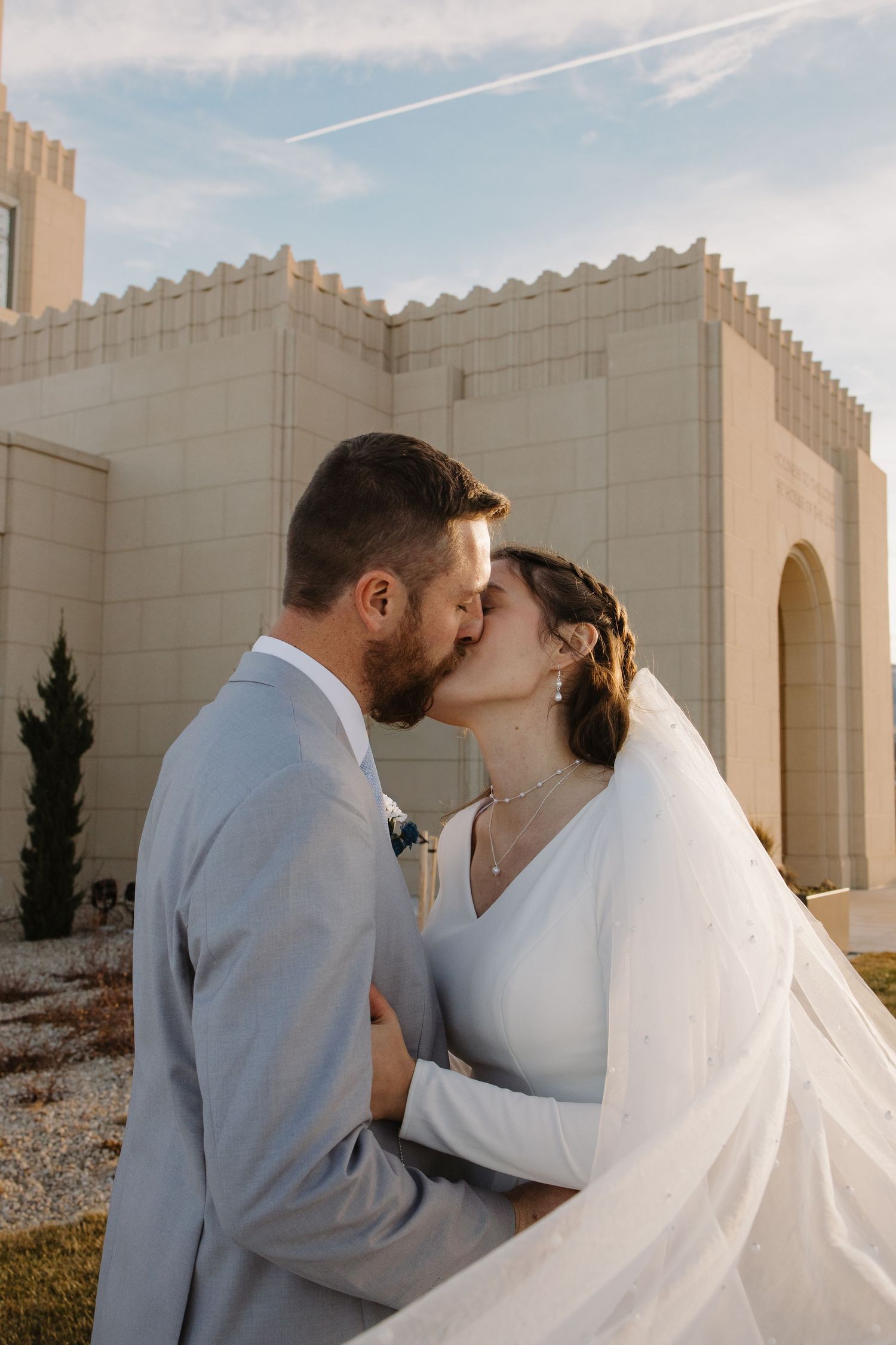 Newlyweds share a kiss outside a white temple building under a blue sky on their wedding day.