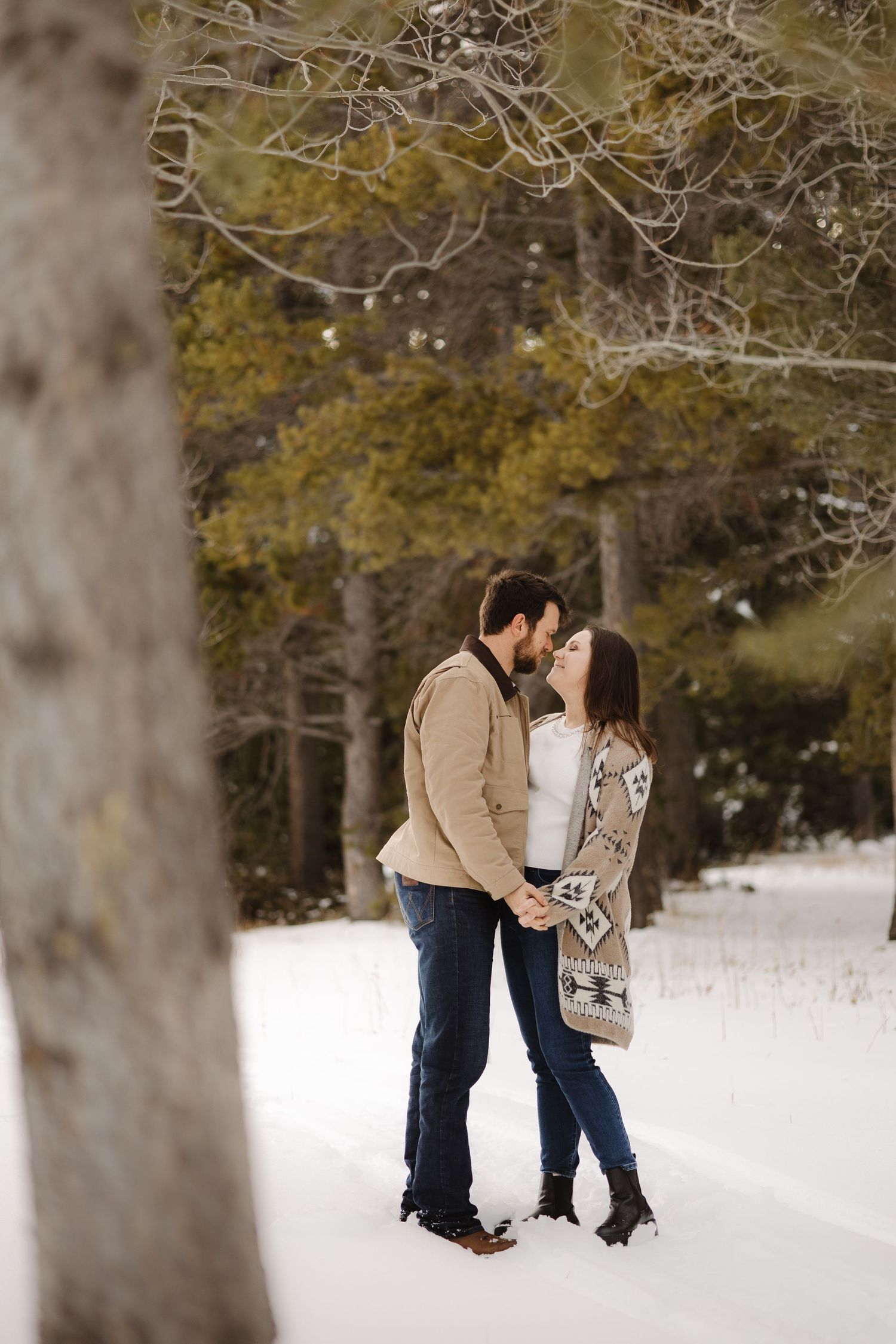 Couple shares a romantic kiss while holding hands in a snow-covered forest setting.