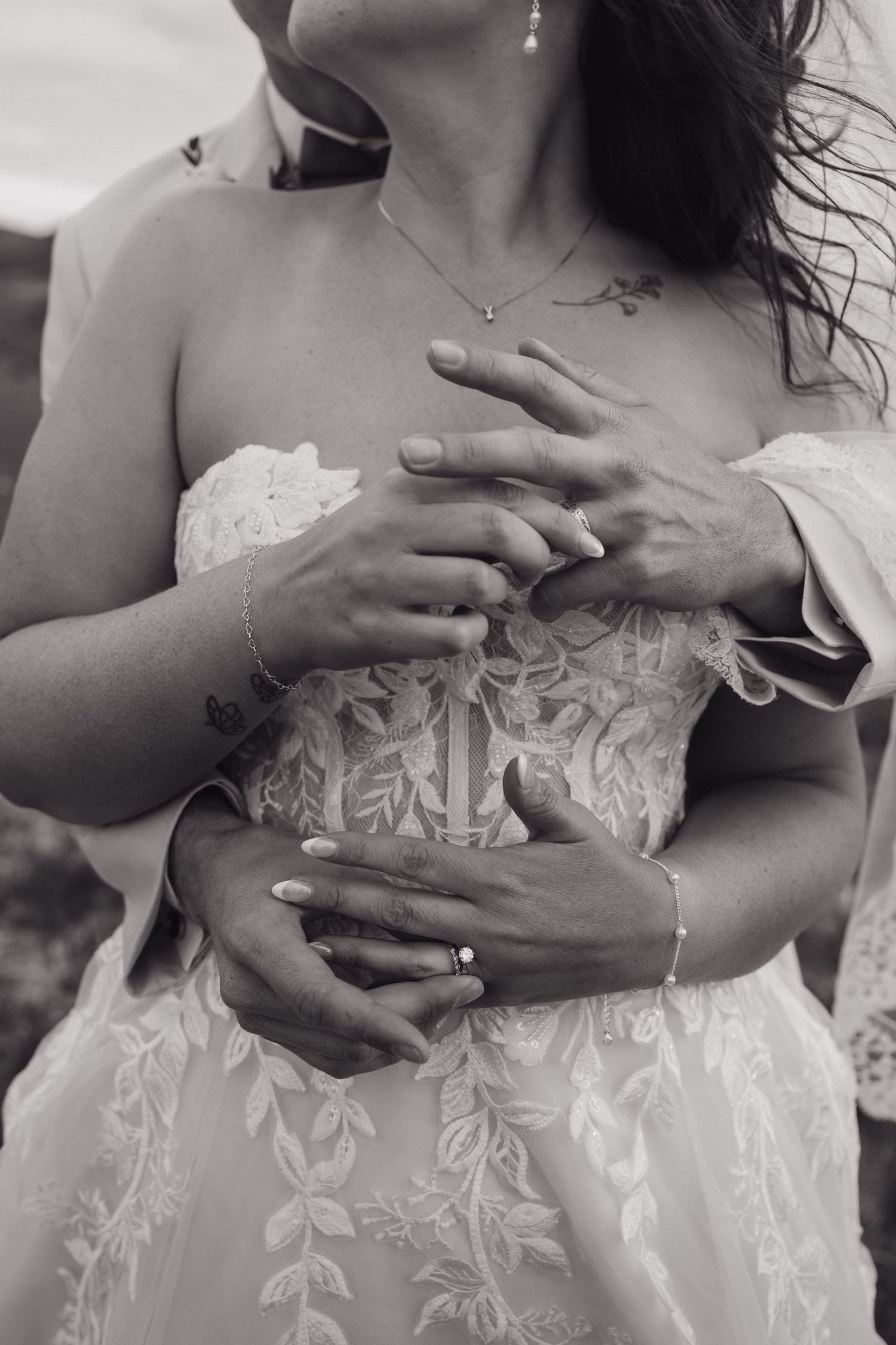 Black and white photo of bride and groom hands embracing, showing wedding rings and lace bridal gown.
