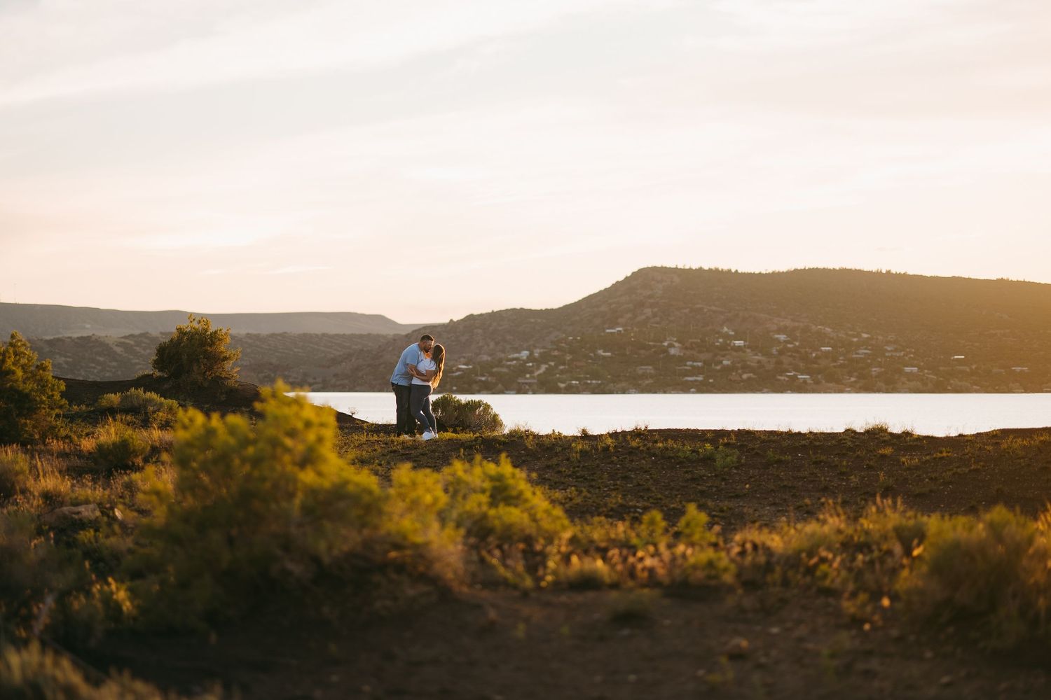 Couple embracing near a scenic lake at golden sunset with rolling hills in the background.