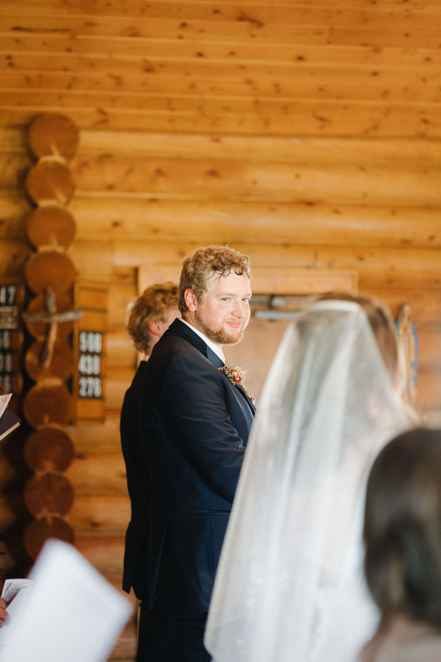 Groom in navy suit smiles at bride walking down the aisle in a rustic log cabin church.