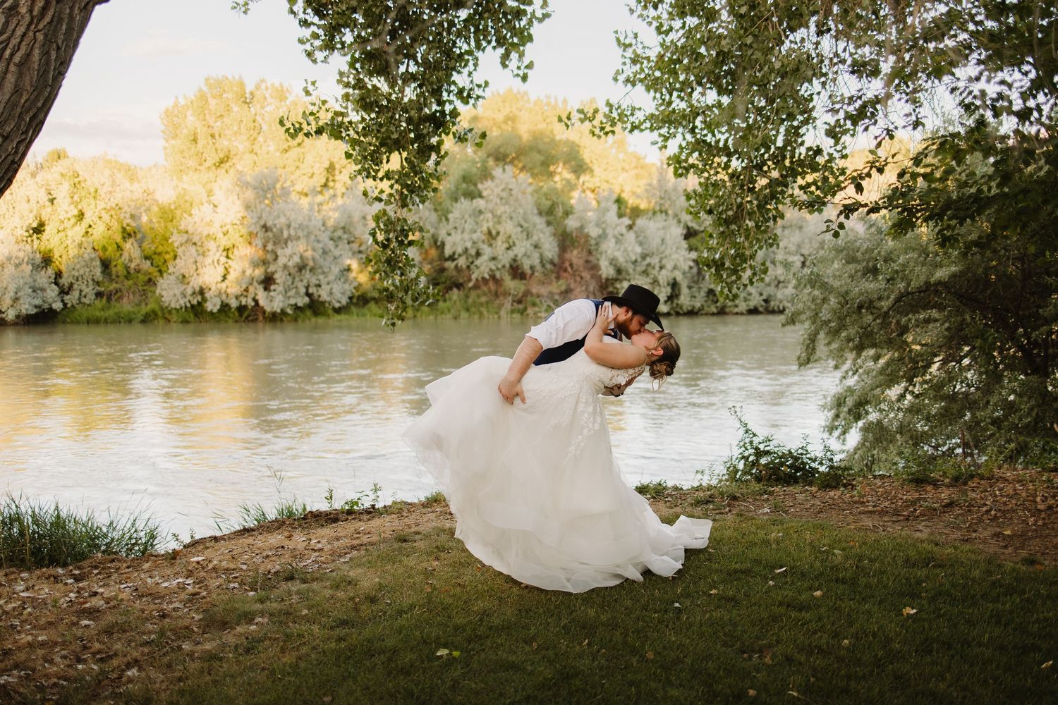 Groom dips bride in flowing white gown by a serene riverside surrounded by lush green trees.