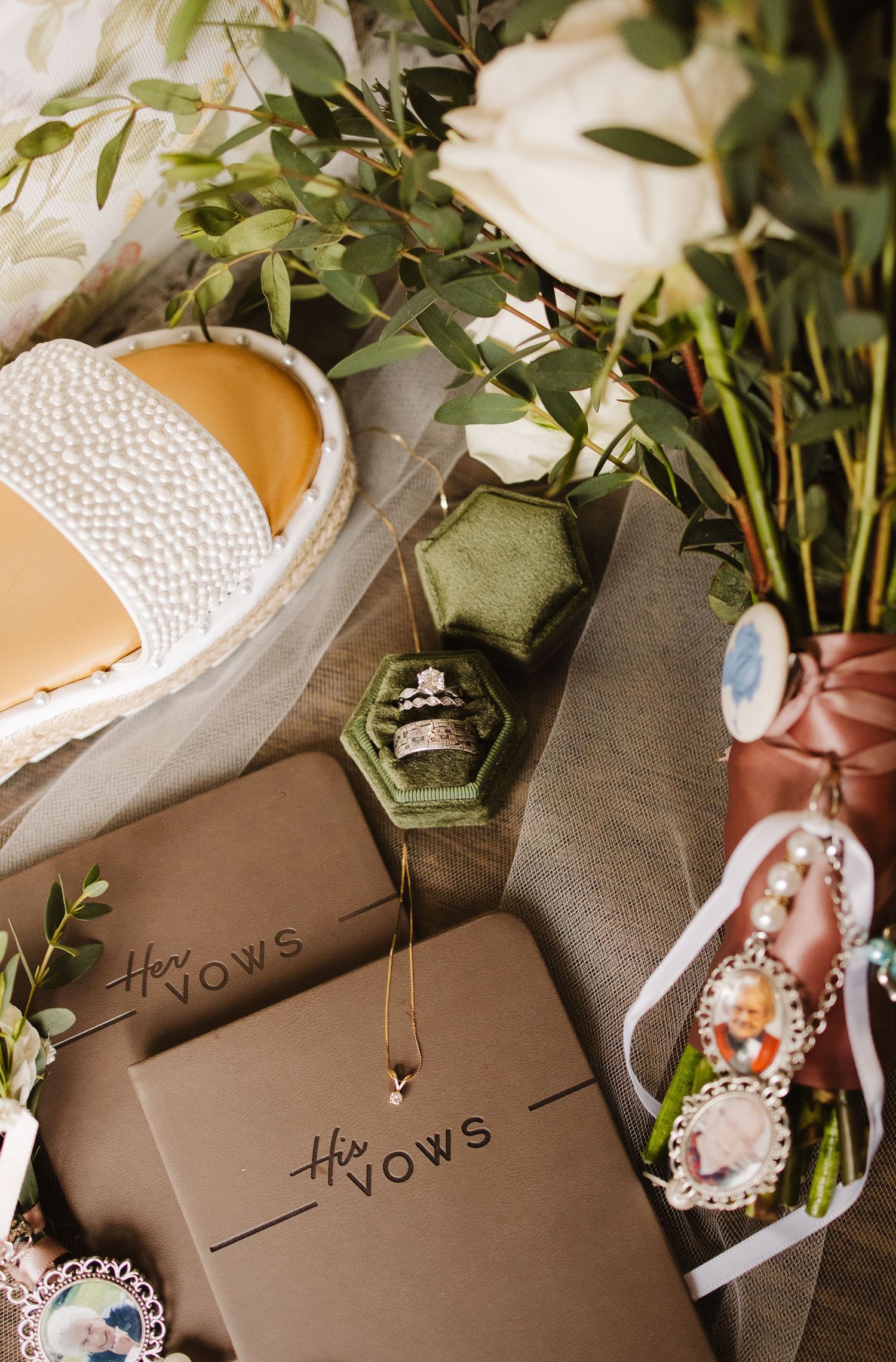 Wedding flat lay featuring vow books, green velvet ring box, bridal bouquet, and memorial charms.