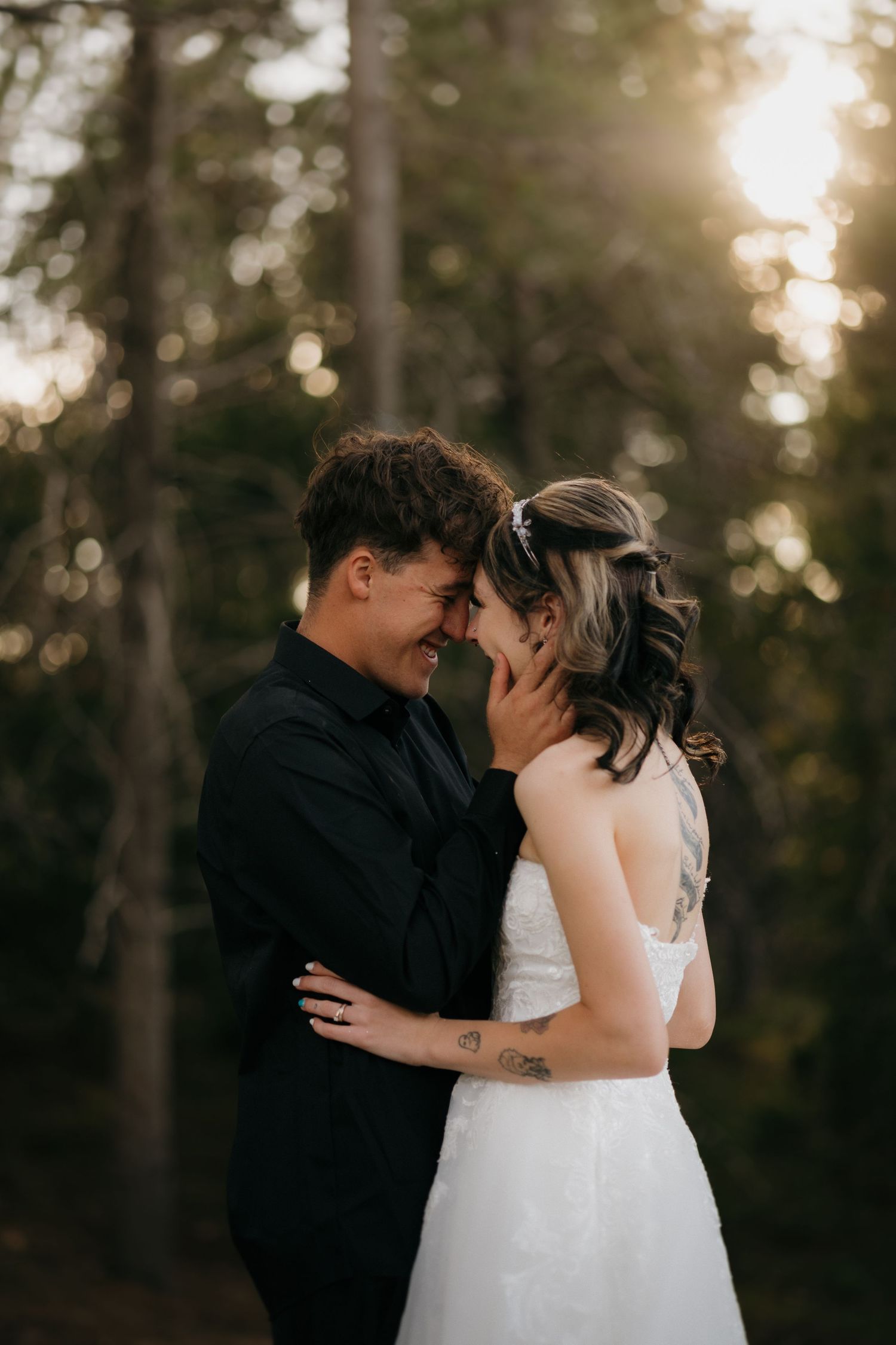 Bride and groom touching foreheads tenderly in a sunlit forest during golden hour.